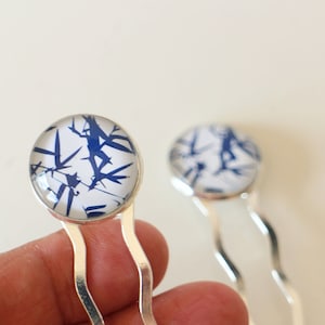 Puede incluir: Dos pinzas para el pelo plateadas con detalles redondos de vidrio con patrones azules y blancos. El patrón se asemeja al bambú.