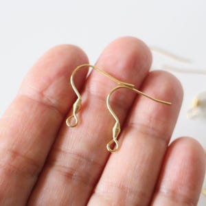 May include: Two gold-toned earring hooks, also known as ear wires, are held in a hand. The hooks are made of metal and have a small loop at the top for attaching earrings.