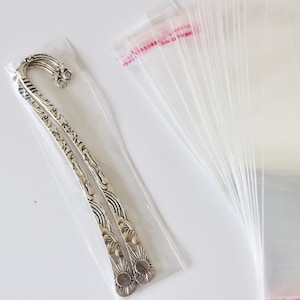 Puede incluir: Un marcapáginas decorativo de metal plateado con un diseño ornamentado está empaquetado en una funda de plástico transparente. También se ve una pila de bolsas de celofán transparentes con una parte superior sellada rosa y roja.