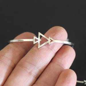 May include: A silver cuff bracelet with a geometric arrow design. The bracelet features three connected triangles at the centre. The open design allows for easy wear. The bracelet is shown against a dark background.