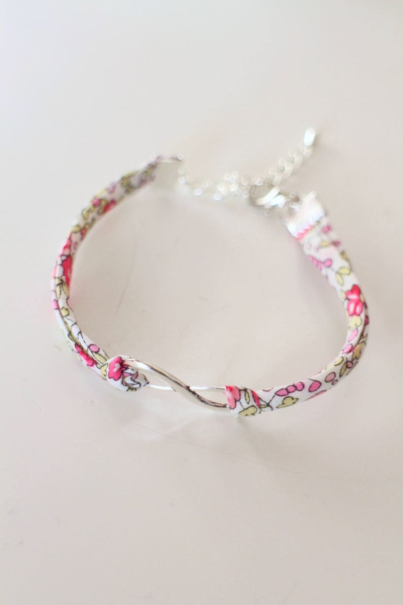 Puede incluir: Un brazalete de tela blanca con un patr&oacute;n floral rosa y un cierre de plata en forma de infinito.