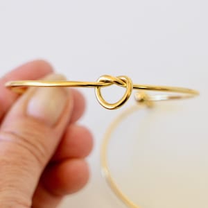 Peut inclure: Un bracelet jonc doré avec un motif de nœud en forme de cœur. Le bracelet est tenu par une main, avec un autre bracelet similaire visible en arrière-plan. Le bijou est posé sur un fond blanc.