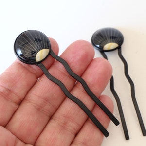 Puede incluir: Dos palos de pelo negros con extremos redondos decorativos. Los extremos son negros con un diseño de sol radiante blanco y dorado.