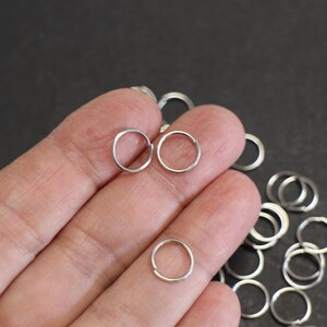 Puede incluir: Una colección de pequeños pendientes de aro plateados. Varios pendientes están esparcidos sobre una superficie oscura, algunos se sostienen en una mano. Los pendientes son simples, circulares y parecen ser de metal.