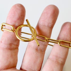 Peut inclure: Un bracelet doré avec un fermoir à bascule. Le bracelet présente un motif de maillons de chaîne avec des maillons rectangulaires. Le fermoir est un anneau circulaire avec une barre droite qui le traverse.