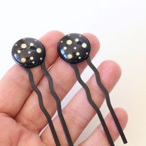 Puede incluir: Dos horquillas negras con tapas redondas de lunares dorados. Las horquillas se sostienen en una mano.