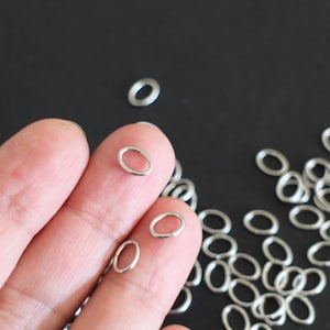 Puede incluir: Primer plano de un montón de anillos de salto plateados. Los anillos son de forma ovalada y tienen un diámetro de aproximadamente 5 mm. Los anillos están dispersos sobre una superficie negra.