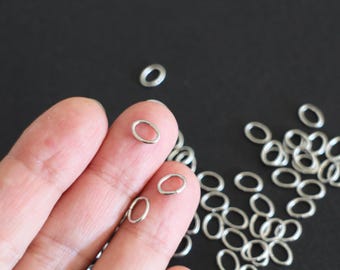 Puede incluir: Primer plano de un montón de anillos de salto plateados. Los anillos son de forma ovalada y tienen un diámetro de aproximadamente 5 mm. Los anillos están dispersos sobre una superficie negra.