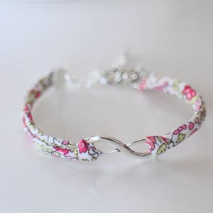 Puede incluir: Un brazalete de tela blanca con un patr&oacute;n floral rosa y un dije de infinito plateado.