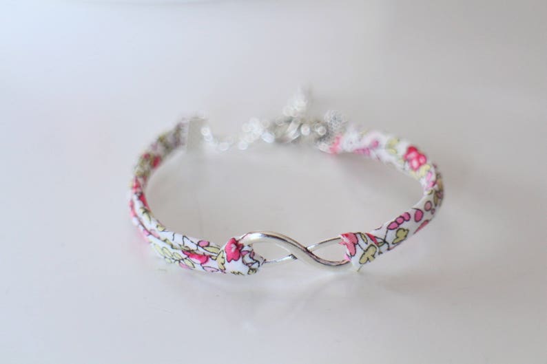 Puede incluir: Un brazalete de tela blanca con un patr&oacute;n floral rosa y un dije de infinito plateado.