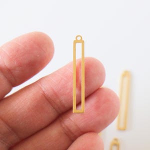 Puede incluir: Un colgante rectangular dorado con un centro hueco y un pequeño lazo en la parte superior. El colgante se sostiene en una mano sobre un fondo blanco. Hay colgantes adicionales visibles en el fondo.