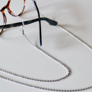 Op de afbeelding: Een zilveren kettingbrilhouder met een sluiting aan elk uiteinde. De ketting is gemaakt van kleine, met elkaar verbonden ovale vormen.