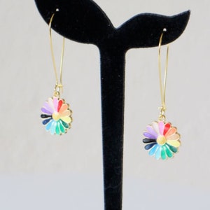 Op de afbeelding: Een paar gouden oorhangers met regenboogkleurige madeliefjescharms. De oorbellen worden getoond op een zwarte oorbelstandaard.