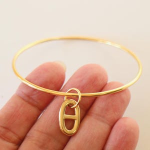 Op de afbeelding: Een goudkleurige armband met een bedel. De bedel heeft een ovale vorm en is goudkleurig, met een horizontale balk in het midden. De armband wordt in een hand gehouden tegen een witte achtergrond. De armband heeft een diameter van ongeveer 6,35 cm.