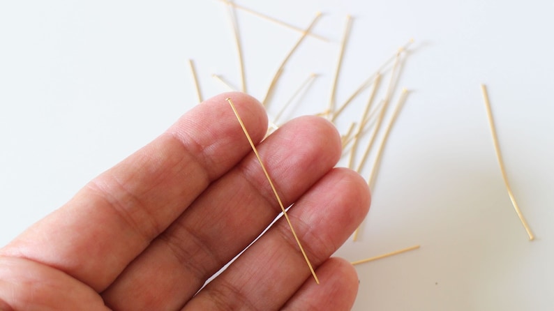 Pu&ograve; includere: Primo piano di una mano che tiene uno spillo dritto dorato. Lo spillo &egrave; lungo circa 2,5 cm ed &egrave; circondato da altri spilli su una superficie bianca.