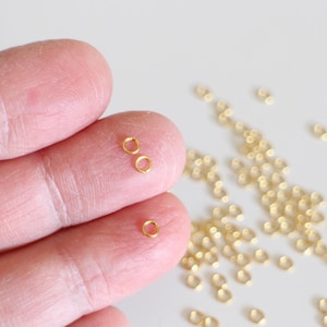 Op de afbeelding: Een close-up van een hand die twee kleine gouden springringen vasthoudt. Een stapel gouden springringen ligt verspreid op een wit oppervlak.