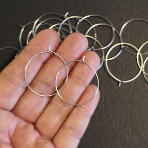 Peut inclure: Une collection de composants de boucles d'oreilles créoles argentées. Plusieurs créoles ouvertes sont visibles, dont deux sont tenues dans une main. Les boucles d'oreilles sont en fil métallique fin et ont un design simple et minimaliste. Le fond est de couleur sombre.