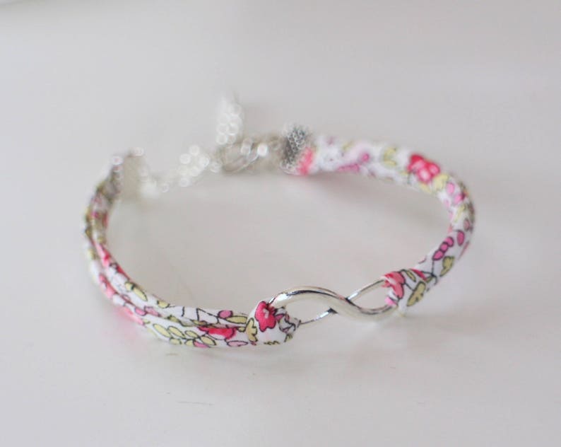Puede incluir: Un brazalete de tela blanca con un estampado floral rosa y amarillo y un cierre de s&iacute;mbolo de infinito plateado.