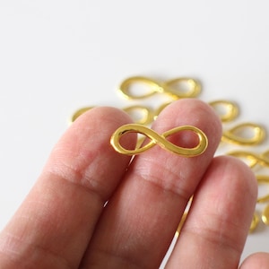 Könnte beinhalten: Ein goldener Unendlichkeits-Anhänger aus der Nähe. Der Anhänger wird von einer Hand gehalten, und im Hintergrund befinden sich mehrere andere Anhänger.