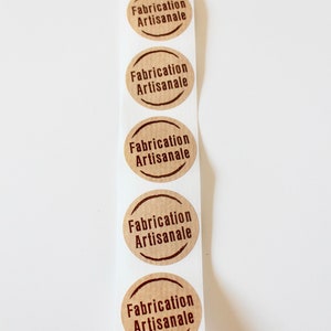 May include: A roll of brown paper stickers with the text "Fabrication Artisanale" printed in dark brown on each sticker.