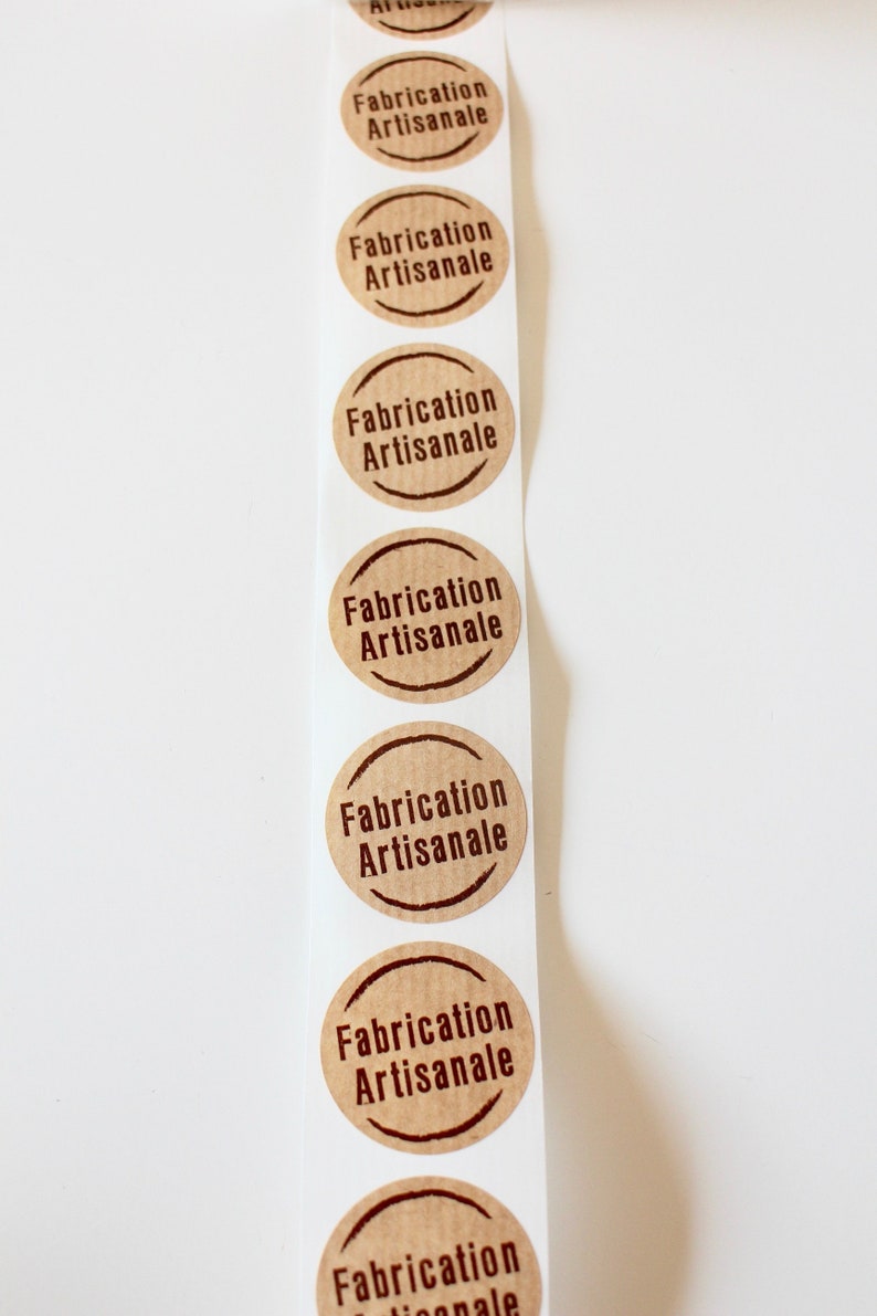 May include: A roll of brown paper stickers with the text "Fabrication Artisanale" printed in dark brown on each sticker.