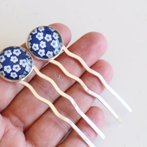 Puede incluir: Dos pinzas para el pelo plateadas con cabujones redondos con estampado floral azul y blanco.