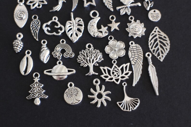 May include: A collection of silver charm pendants featuring various nature-inspired designs, including leaves, trees, flowers, a seashell, a rainbow, a star, a sun, a moon, a feather, a pine cone, a cloud, a planet, a lotus flower, a tree of life, a ginkgo leaf, and an angel wing.