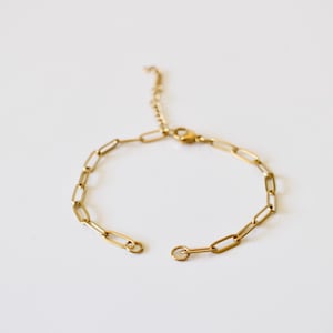 Può includere: Un braccialetto a catena color oro con chiusura aperta. Il braccialetto presenta una serie di maglie rettangolari collegate tra loro. La catena regolabile si estende dalla catena principale. Il braccialetto è esposto su una superficie bianca.