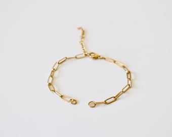 Puede incluir: Una pulsera de cadena dorada con un cierre abierto. La pulsera presenta una serie de eslabones rectangulares conectados entre sí. La cadena ajustable se extiende desde la cadena principal. La pulsera se muestra sobre una superficie blanca.
