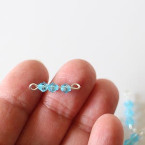Pode incluir: Um close-up de uma mão segurando um fio de prata com quatro contas de cristal azul claro presas.