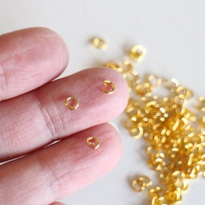 Puede incluir: Primer plano de una mano sosteniendo dos anillos de salto dorados. Un mont&oacute;n de anillos de salto dorados se dispersa sobre una superficie blanca.