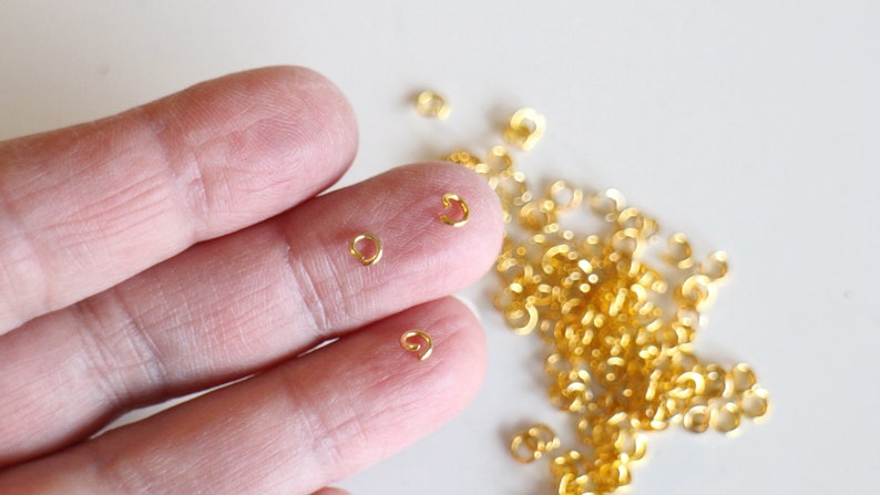 Puede incluir: Primer plano de una mano sosteniendo dos anillos de salto dorados. Un mont&oacute;n de anillos de salto dorados se dispersa sobre una superficie blanca.