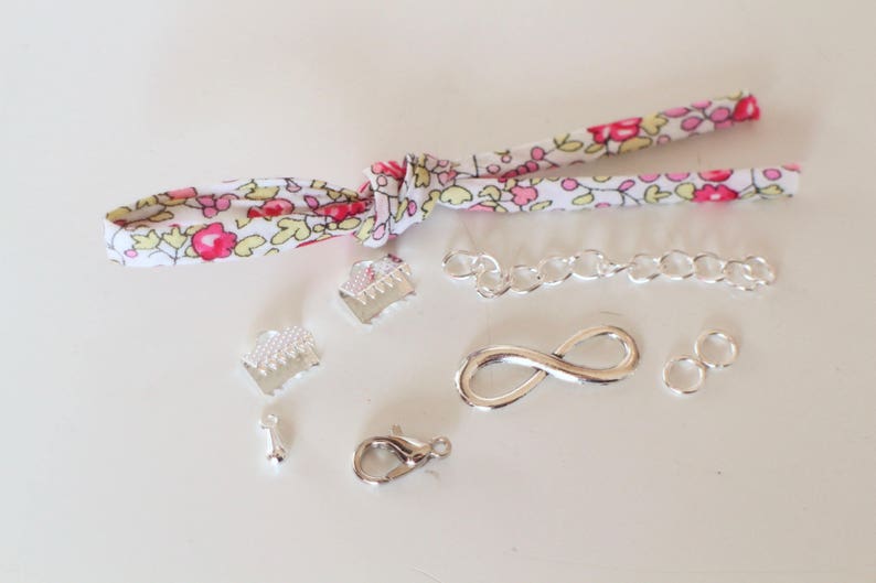 Puede incluir: Una colecci&oacute;n de suministros de fabricaci&oacute;n de joyas plateados, que incluyen una cadena, un amuleto de infinito, anillos de salto y cierres. Tambi&eacute;n hay un trozo de tela con un patr&oacute;n floral.