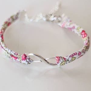 Puede incluir: Un brazalete de tela blanca con un patr&oacute;n floral rosa y un cierre de plata en forma de infinito.