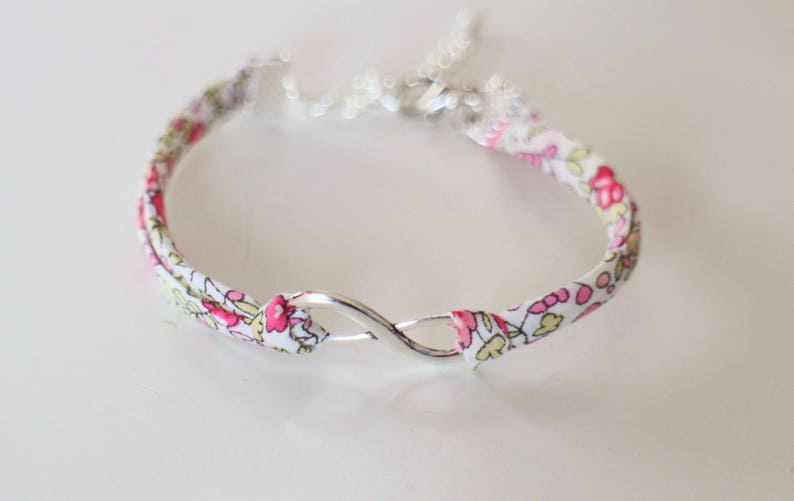Puede incluir: Un brazalete de tela blanca con un patr&oacute;n floral rosa y un cierre de plata en forma de infinito.