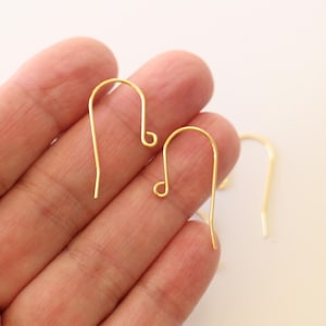 May include: Two gold-toned earring hooks, also known as French hooks, are shown in a hand. The hooks are made of metal and have a simple design.