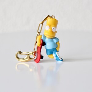 Może przedstawiać: Żółta figurka Barta Simpsona z czerwoną deskorolką, niebieskimi szortami i jasnoniebieską koszulą. Brelok ma złote zapięcie i łańcuszek. Figurka opiera się o deskorolkę.