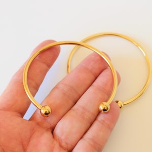 Peut inclure: Deux bracelets dorés ouverts. L'un est tenu dans une main, l'autre repose sur une surface blanche. Chaque bracelet a une petite boule ronde à chaque extrémité. Les bijoux sont simples et élégants.