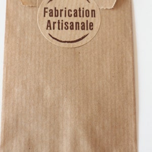 May include: A brown paper bag with a round, brown label that says "Fabrication Artisanale" in brown text.
