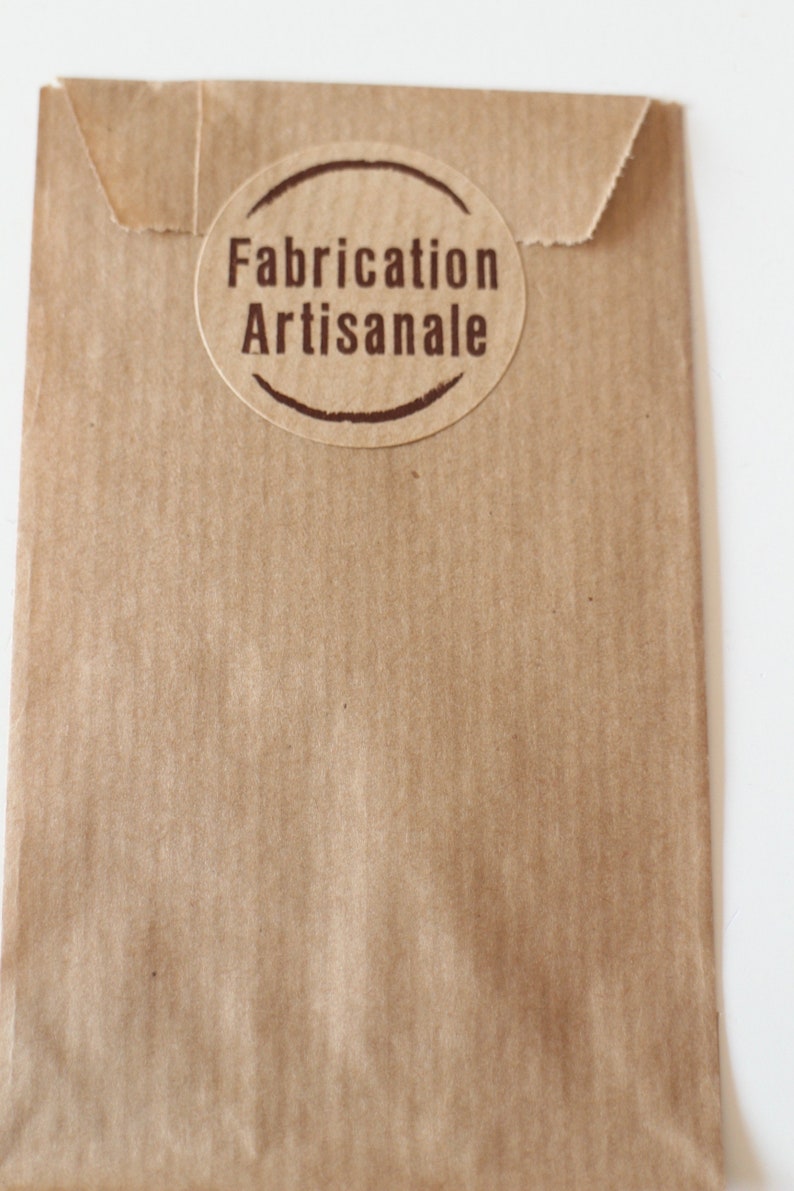 May include: A brown paper bag with a round, brown label that says "Fabrication Artisanale" in brown text.