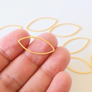 Puede incluir: Hallazgos de joyería dorados en forma de hoja con marco abierto. Varios de estos delicados componentes de forma ovalada están dispersos sobre una superficie blanca, uno de ellos sostenido en una mano. Estos hallazgos son ideales para crear pendientes, colgantes u otras joyas.