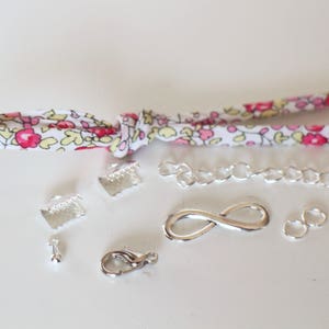 Puede incluir: Una colecci&oacute;n de hallazgos de joyer&iacute;a plateados, que incluyen una cadena, un amuleto de infinito, cierres y anillos de salto. Los hallazgos est&aacute;n dispuestos sobre una superficie blanca con una cinta de tela con estampado floral.