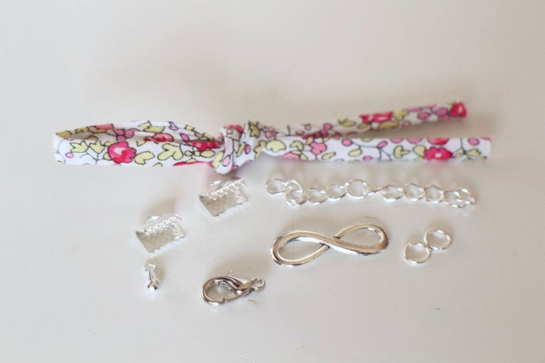 Puede incluir: Una colecci&oacute;n de hallazgos de joyer&iacute;a plateados, que incluyen una cadena, un amuleto de infinito, cierres y anillos de salto. Los hallazgos est&aacute;n dispuestos sobre una superficie blanca con una cinta de tela con estampado floral.