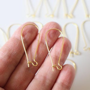 May include: Gold-toned earring hooks, a pair of hooks are held in a hand, with several more hooks scattered on a white surface.