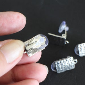 May include: Three silver earring clips with clear rubber backs. The clips are rectangular and have a textured surface.