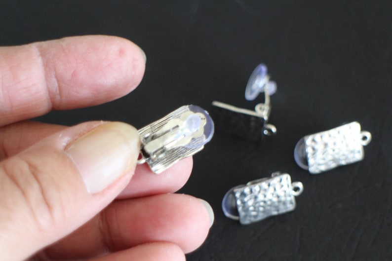 May include: Three silver earring clips with clear rubber backs. The clips are rectangular and have a textured surface.