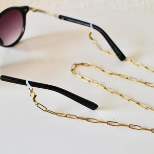 Op de afbeelding: Zwarte zonnebril met verloopglazen en goudkleurige accenten, gecombineerd met een gouden brillenketting. De bril heeft een ronde vorm en de ketting is gemaakt van langwerpige schakels.
