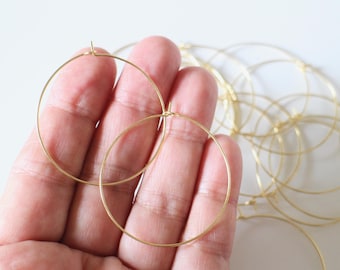 May include: A hand holds two gold-colored hoop earrings. The earrings are thin and circular, with a simple design. Other similar earrings are visible in the background, suggesting a collection or set. The background is a plain white surface.