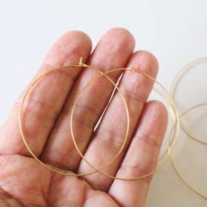 May include: A close-up of several gold-colored hoop earrings. Two large hoops are held in a hand, with several other hoops visible in the background. The earrings are thin and delicate, with a simple design.