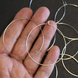 Può includere: Una collezione di orecchini a cerchio color argento. Gli orecchini sono sottili e circolari, con un design semplice. Sono visibili diversi cerchi, alcuni sovrapposti. Gli orecchini sono esposti su uno sfondo scuro.